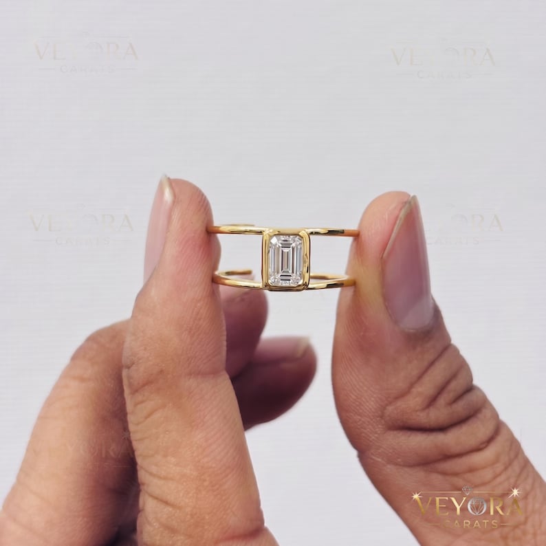 May include: A gold ring featuring a central emerald-cut clear gemstone held in a bezel setting. The band consists of two parallel gold wires that connect at the sides to the central stone setting, creating an open, split-shank design.