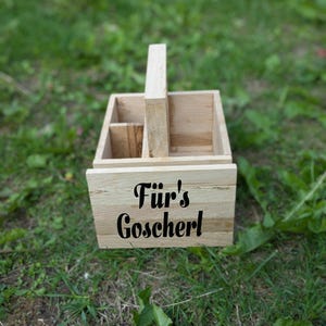 Op de afbeelding: Een lichtgekleurde houten caddy met een handvat, met compartimenten en de tekst "F&uuml;r's Goscherl" in zwart schrift. De caddy staat op een bed van groen gras.