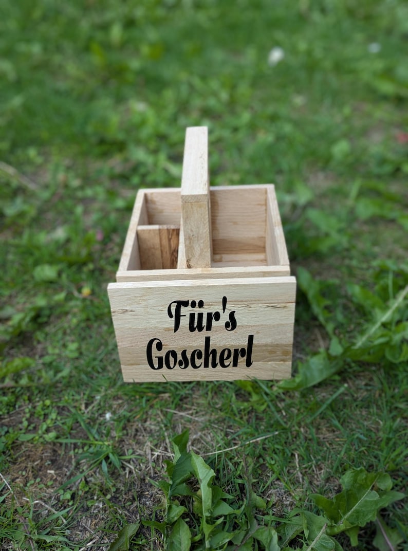 Op de afbeelding: Een lichtgekleurde houten caddy met een handvat, met compartimenten en de tekst "F&uuml;r's Goscherl" in zwart schrift. De caddy staat op een bed van groen gras.