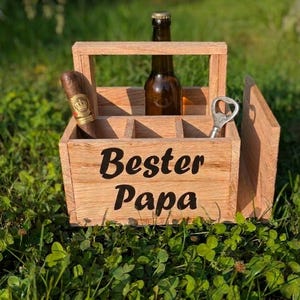 Op de afbeelding: Houten bierkrat met de woorden "Bester Papa" in zwart schrift. De krat bevat een bierfles, een sigaar en een flesopener. De krat staat op een bed van groen gras.