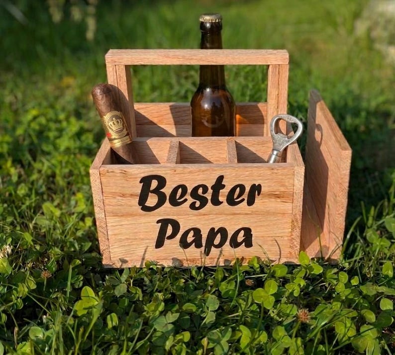Op de afbeelding: Houten bierkrat met de woorden "Bester Papa" in zwart schrift. De krat bevat een bierfles, een sigaar en een flesopener. De krat staat op een bed van groen gras.
