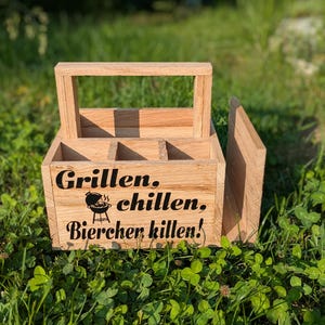Op de afbeelding: Een houten bierkrat met een handvat en vakken. De krat heeft de woorden "Grillen, chillen, Bierchen killen!" in zwarte letters, samen met een afbeelding van een grill. De krat is gemaakt van lichtgekleurd hout en staat op gras.