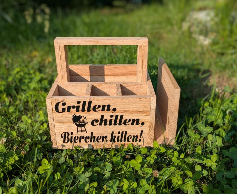 Op de afbeelding: Een houten bierkrat met een handvat en vakken. De krat heeft de woorden "Grillen, chillen, Bierchen killen!" in zwarte letters, samen met een afbeelding van een grill. De krat is gemaakt van lichtgekleurd hout en staat op gras.