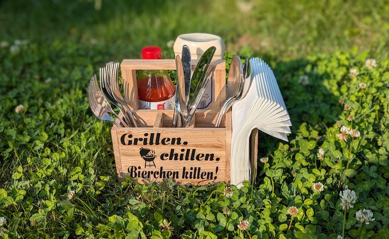 Op de afbeelding: Een houten bestekhouder met de woorden "Grillen. chillen. Bierchen killen!" en een barbecue-afbeelding. Het bevat vorken, messen, lepels, servetten, ketchup en een kruidenpotje. De houder staat in groen gras.