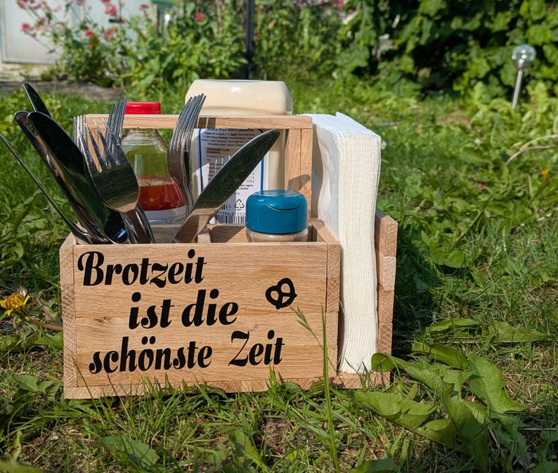 Op de afbeelding: Een houten caddy gevuld met bestek, kruiden en servetten, perfect voor buiten dineren. De caddy heeft de woorden "Brotzeit ist die sch&ouml;nste Zeit" en een pretzelsymbool. Inclusief een pot met een blauw deksel.