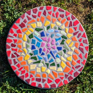 Puede incluir: Una losa de jardín redonda de mosaico con un borde gris y un diseño abstracto colorido. El mosaico presenta una variedad de colores, incluyendo rojo, naranja, amarillo, verde, azul y morado, dispuestos en un patrón circular. La piedra está sobre hierba verde.
