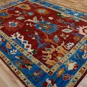 May include: A rectangular area rug with a deep red field and a vibrant blue border. The rug features intricate floral and geometric patterns in shades of blue, gold, and white. The rug has a fringed edge and is placed on a light wood floor.