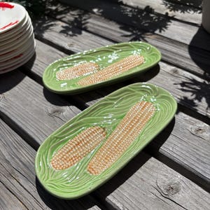 May include: Two ceramic serving platters shaped like corn on the cob. The platters are light green with a textured leaf design around the edges. The corn kernels are tan and detailed. The platters are on a weathered wooden surface.
