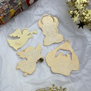 May include: Four gold-colored Christmas ornaments. One angel plays a trumpet, another kneels, and a third holds a bell. The fourth ornament depicts a nativity scene. Ornaments are on white paper with gold star garland.