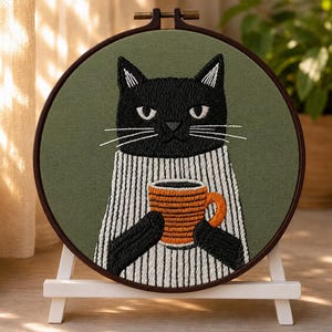 May include: A circular embroidery hoop containing a fabric panel with a stitched black cat wearing a white jumper with black vertical stripes. The cat holds an orange mug with horizontal lines. The background fabric is a solid olive green colour.