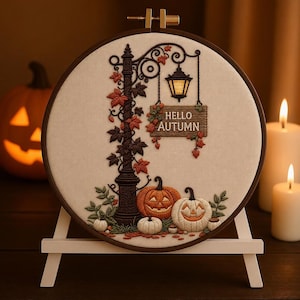 Puede incluir: Un bastidor de bordado con un diseño de farola marrón oscuro con hojas de otoño, un letrero colgante que dice HELLO AUTUMN y tres calabazas talladas en la base, bordado sobre tela color crema y exhibido sobre un soporte de color blanco.
