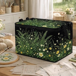 Puede incluir: Una caja de almacenamiento rectangular negra con un estampado botánico de helechos verdes y flores silvestres amarillas. El contenedor cuenta con una tapa con cremallera y asas de tela negra en los laterales para organizar objetos.