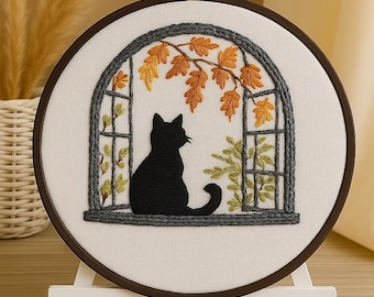 Pode incluir: Arte bordada em um bastidor de madeira. O design mostra um gato preto olhando por uma janela com moldura cinza. A janela apresenta folhas de outono em laranja e amarelo. O bastidor está sobre um suporte branco.