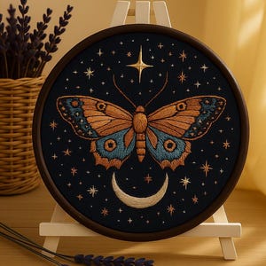 Puede incluir: Obra de arte bordada redonda que representa una mariposa con alas naranjas y azules sobre un fondo azul oscuro estrellado. El diseño incluye una luna creciente y una estrella. La obra se exhibe en un caballete de madera.