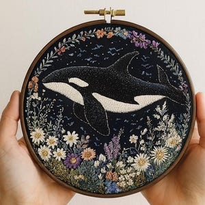 May include: Embroidered artwork featuring a black and white orca swimming in a dark blue sea, surrounded by colorful flowers. The orca is the focal point, with detailed stitching. The artwork is framed in a round wooden hoop.