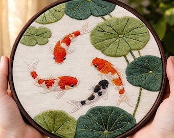 May include: Embroidered artwork featuring koi fish and lily pads. The design includes orange, red, and black koi fish swimming among green lily pads. The artwork is displayed in a round wooden embroidery hoop.