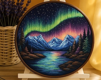 Pode incluir: Um bastidor de bordado redondo com uma cena de paisagem da Aurora Boreal. A obra de arte retrata um lago, montanhas e árvores sob uma exibição vibrante de luzes verdes, roxas e rosas. O bastidor é exibido em um cavalete de madeira.