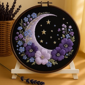 Può includere: Ricamo su telaio raffigurante una luna crescente e stelle su sfondo nero, circondato da fiori viola e blu. Il ricamo è incorniciato in un telaio di legno e presentato su un cavalletto bianco.