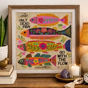 May include: A framed artwork featuring four colorful, embroidered fish with floral designs. The text "ONLY DEAD FISH GO WITH THE FLOW" is displayed. The artwork is set on a neutral background, with the brand name "natural life" at the bottom.