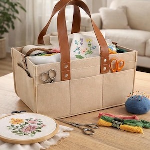 May include: A beige felt craft organizer with brown leather handles, filled with embroidery supplies. Includes scissors, thread, a blue pin cushion, and an embroidery hoop with a floral design. The organizer sits on a wooden table.