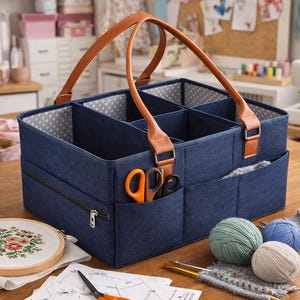 Può includere: Un organizer per artigianato in tessuto blu con più scomparti e un manico in pelle marrone. L'organizer ha una tasca con cerniera e diverse tasche esterne. Forbici arancioni e nere sono in una tasca. Forniture per maglieria e un telaio per ricamo sono nelle vicinanze.