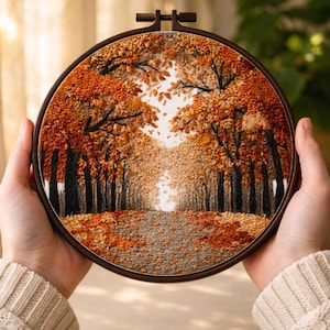 Könnte beinhalten: Ein runder Stickrahmen mit einer detaillierten Herbstszene. Das Kunstwerk zeigt einen von Bäumen gesäumten Weg mit leuchtend orangefarbenem und braunem Laub. Die Stickerei wird von Händen gehalten, die die Textur und Farben zeigen.