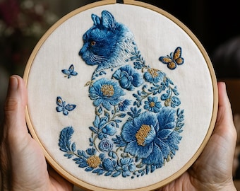 Pode incluir: Uma obra de arte bordada com a silhueta de um gato azul adornada com padrões florais e borboletas. O gato está sobre um tecido creme, emoldurado num bastidor de madeira. O bordado apresenta pontos detalhados e uma mistura de tons azuis e dourados.