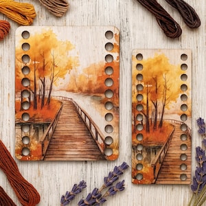 May include: Two wooden embroidery hoops with a landscape painting of a bridge over a river in autumn colors. The hoops have multiple holes for stitching. Embroidery floss and lavender sprigs are arranged around the hoops.
