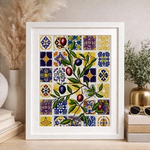 Puede incluir: Un bordado en punto de cruz enmarcado que presenta una rama de olivo central con aceitunas de color púrpura oscuro y hojas verdes, sobre un fondo de azulejos cuadrados coloridos y repetitivos en tonos azul, amarillo y púrpura en un marco blanco.