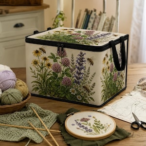 May include: A beige storage tote with a black zipper and handles, adorned with a botanical print of wildflowers and bees. The tote is on a wooden table with knitting supplies and an embroidery hoop.