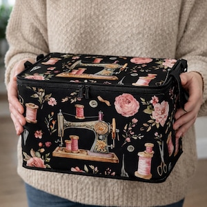 Könnte beinhalten: Ein rechteckiger schwarzer Aufbewahrungskoffer mit einem wiederkehrenden Muster aus alten Nähmaschinen, rosa Garnrollen, Blüten und silbernen Scheren. Die Tasche verfügt über einen Reißverschluss oben und schwarze Tragegriffe an den Seiten.