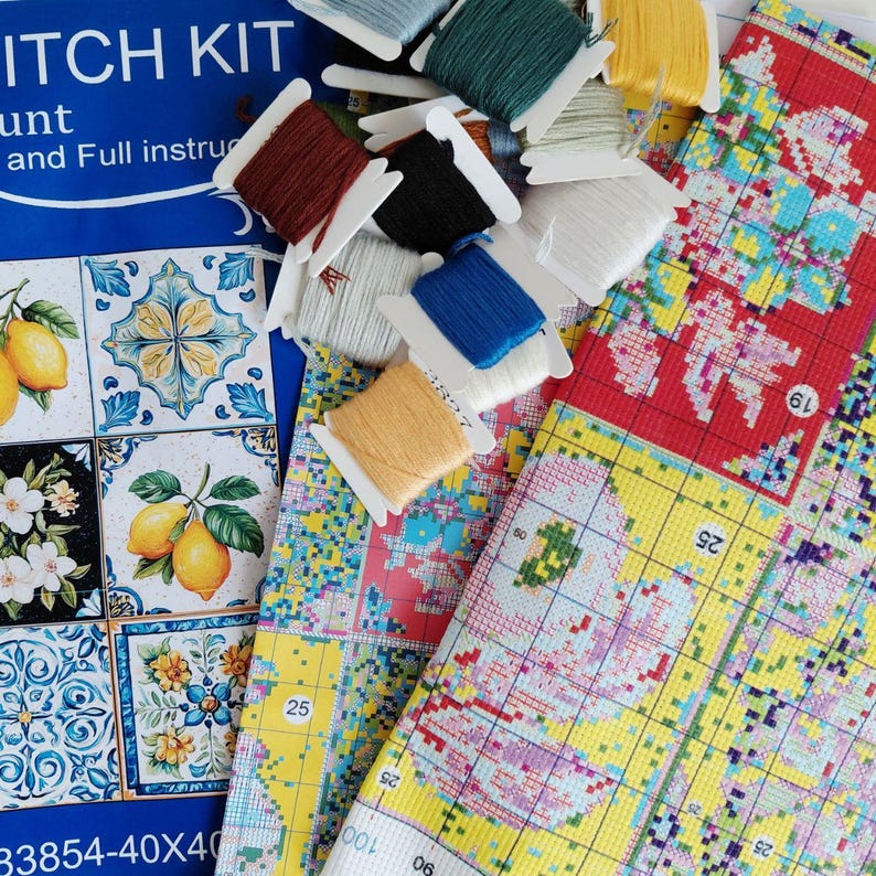 Puede incluir: Un kit de punto de cruz que incluye una hoja de instrucciones azul con patrones de azulejos de limones y flores, una tela cuadriculada con un dise&ntilde;o impreso y varios carretes de hilo de bordar en amarillo, azul, blanco, marr&oacute;n y negro.
