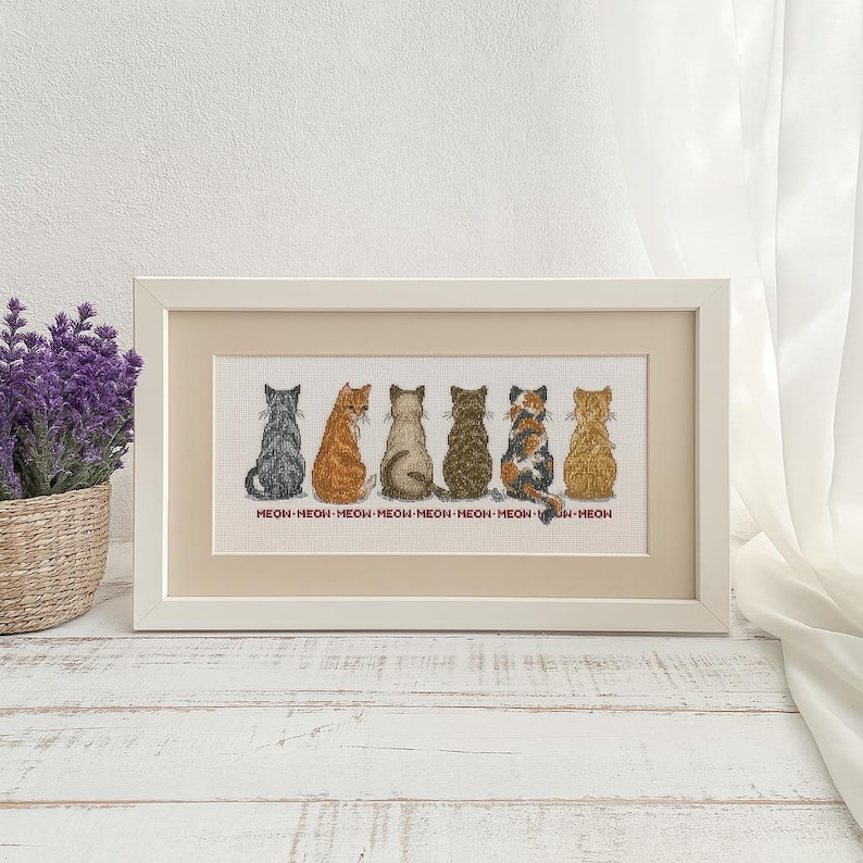 May include: A framed cross-stitch featuring the backs of seven cats in various colors, including gray, orange, and calico. The text "MEOW" is repeated below each cat. The frame is white, and the background is a neutral color.