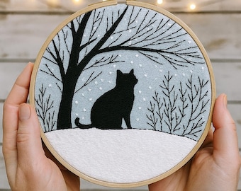 Op de afbeelding: Een ronde borduurring met een zwart katten silhouet in een winterlandschap. De kat zit op een besneeuwde heuvel onder een kale boom en vallende sneeuw. De achtergrond is lichtblauwe stof, vastgehouden door handen.