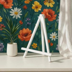 Op de afbeelding: Een klein, wit houten ezel staat op een wit oppervlak, tegen een achtergrond van bloemenbehang met oranje, gele en witte bloemen. Een plant in een pot met groene bladeren staat links. Rechts is een wit gordijn.