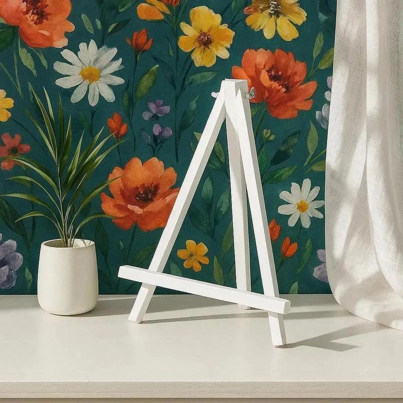 Op de afbeelding: Een klein, wit houten ezel staat op een wit oppervlak, tegen een achtergrond van bloemenbehang met oranje, gele en witte bloemen. Een plant in een pot met groene bladeren staat links. Rechts is een wit gordijn.