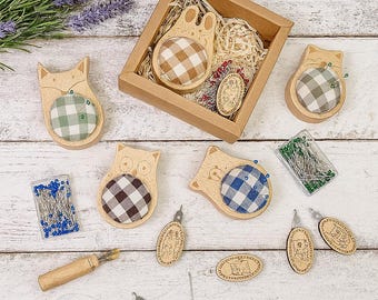 May include: A selection of wooden pin cushions in animal shapes, such as a fox, owl, and bear, with checkered fabric tops. The image also shows small wooden embellishments, pins, and a needle threader, all on a white wooden surface.