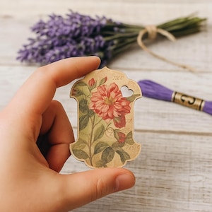 Peut inclure: Une étiquette de bricolage en bois avec un motif floral représentant une rose rouge et des feuilles vertes. L'étiquette a une découpe en forme de cœur et est tenue dans une main. Le fond comprend de la lavande et du fil à broder violet.