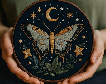 May include: Embroidered artwork featuring a butterfly with detailed wings in shades of gold, gray, and white, set against a dark blue background. The design includes a crescent moon, stars, and floral accents, all within a wooden embroidery hoop.