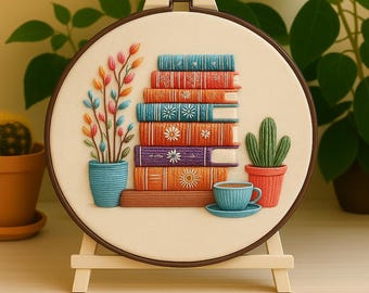 Könnte beinhalten: Gesticktes Hoop-Art mit einem Stapel bunter Bücher, einer Topfpflanze mit farbenfrohem Blumendesign, einem Kaktus und einer Teetasse. Das Kunstwerk wird auf einer kleinen Staffelei präsentiert und schafft ein gemütliches, literarisches Thema.