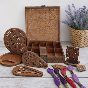 Pode incluir: Um organizador de artesanato de madeira com um design de cruz celta, aberto para revelar vários compartimentos. Peças de madeira adicionais com entalhes intrincados, fios de bordar e um par de tesouras são exibidos. Uma planta de lavanda decorativa em uma cesta tecida está no fundo.