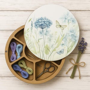 Puede incluir: Un organizador de manualidades redondo de madera con una tapa floral que presenta hortensias azules y mariposas. En el interior, contiene hilo de bordar de varios colores, agujas y tijeras pequeñas. Un ramo de lavanda está atado con hilo.