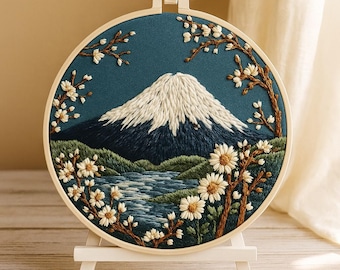 May include: A round, embroidered artwork featuring a mountain landscape. The design includes a snow-capped mountain, a river, and flowering branches. The embroidery is set in a wooden hoop and displayed on a small easel.