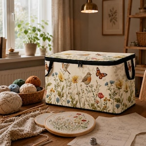 May include: A rectangular storage bin with a cream-colored fabric featuring a botanical print of birds, butterflies, and wildflowers. The bin has black trim, black handles, and a zippered lid. It sits on a wooden table with yarn and embroidery hoops.