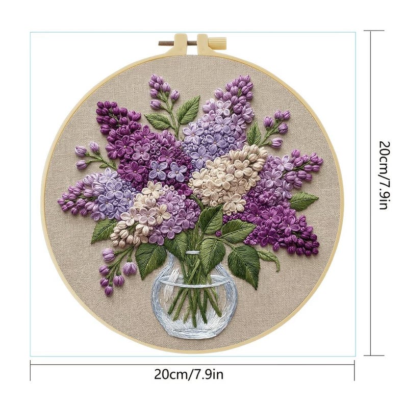 Op de afbeelding: Geborduurde bloemenkunst in een ronde houten hoepel. Het kunstwerk toont een boeket van paarse, lavendelkleurige en witte seringen in een heldere glazen vaas. De borduursels zijn geplaatst tegen een beige stoffen achtergrond. De hoepel heeft een diameter van 20 cm.