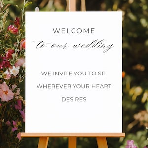 May include: A white wedding sign on a wooden easel, with the words "WELCOME to our wedding" in elegant script. The sign also reads "WE INVITE YOU TO SIT WHEREVER YOUR HEART DESIRES". The background features colorful flowers.