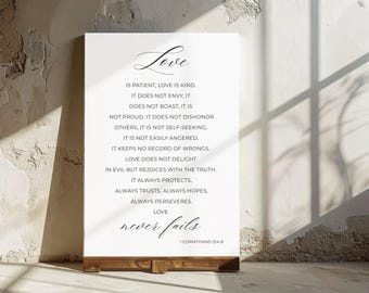 May include: A white canvas sign with the word "Love" in elegant script at the top. The text below lists qualities of love, with the Bible verse 1 Corinthians 13:4-8 at the bottom. The sign is mounted on a wooden base.