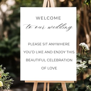 May include: A white sign on a wooden easel with the words "WELCOME to our wedding." The sign also reads "PLEASE SIT ANYWHERE YOU'D LIKE AND ENJOY THIS BEAUTIFUL CELEBRATION OF LOVE."
