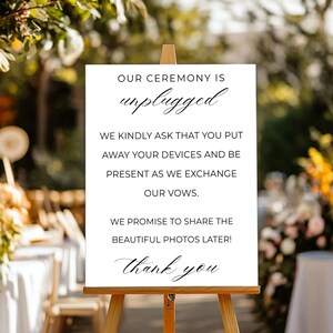 May include: A white sign on a wooden easel with the text "OUR CEREMONY IS unplugged" and additional text requesting guests to put away devices. The sign is set up outdoors with blurred greenery in the background.