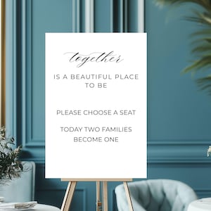 May include: A white sign on a wooden easel with the text "together IS A BEAUTIFUL PLACE TO BE PLEASE CHOOSE A SEAT TODAY TWO FAMILIES BECOME ONE" in black script. The sign is in front of a teal wall with floral arrangements.